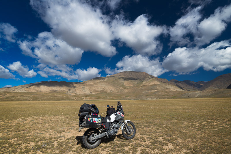 motorcycle parked on a dry-grass field with hills and fluffy clouds behind